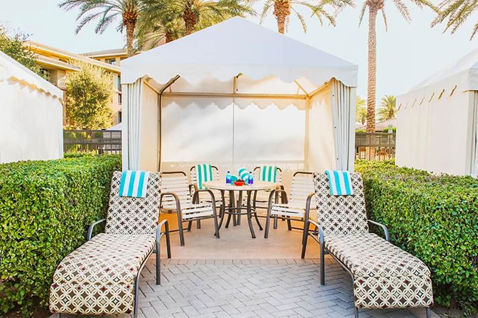 Luxury cabana lounge area with striped chairs, round table, and white canopy tent surrounded by green hedges and palm trees