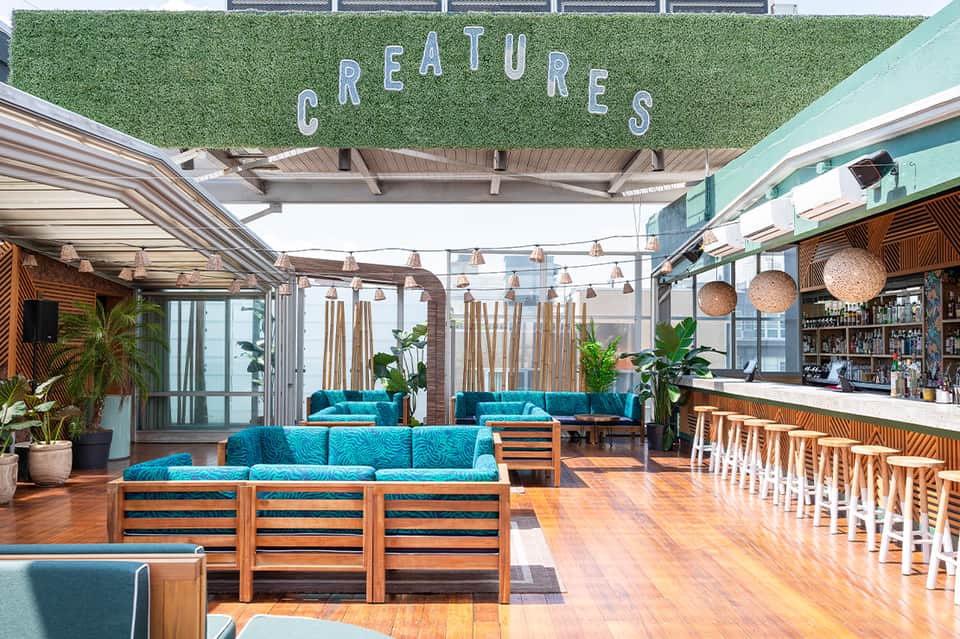 Modern rooftop lounge with turquoise seating, 'Creatures' wall sign, bar area, and hanging lights under geometric ceiling