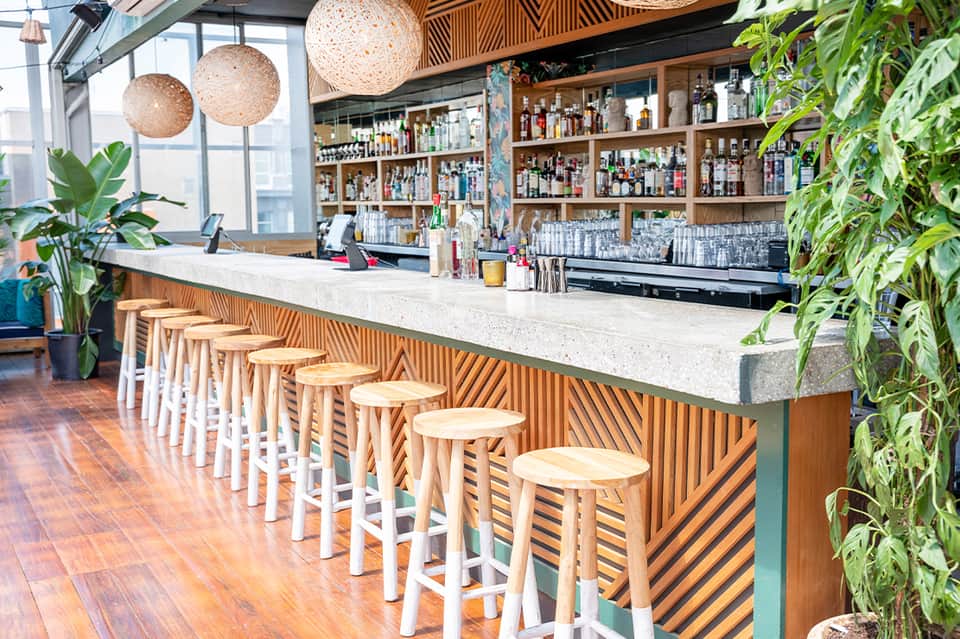 Modern bar with wooden slatted counter, white stools, hanging woven pendant lights, and botanical décor