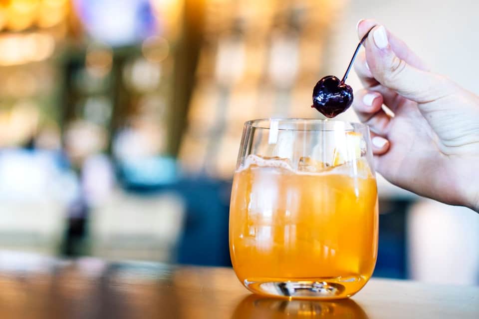 Hand holding cocktail glass with golden liquid and dark cherry garnish on a stick against blurred background