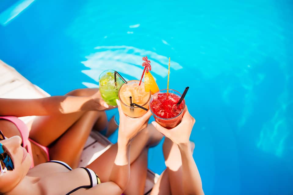 Friends holding tropical cocktails while relaxing by bright blue poolside water