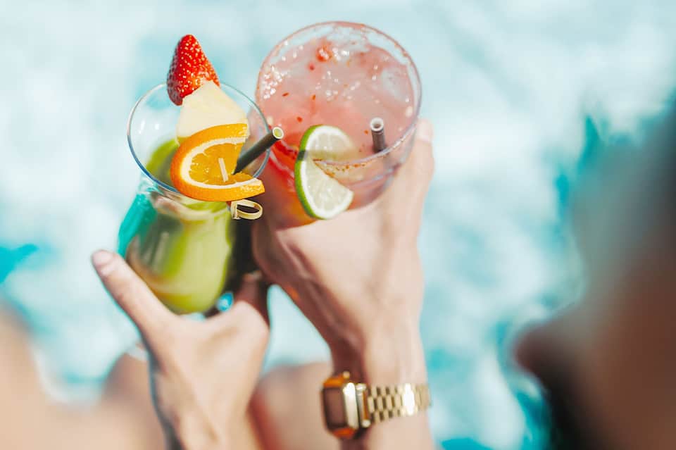 Tropical cocktails held by hands with citrus garnishes and ocean backdrop