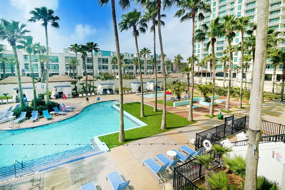 Aerial view of resort pool complex with turquoise water, palm trees, lounge chairs, and beachfront hotel buildings