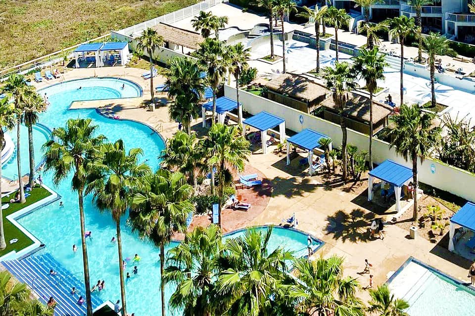 Aerial view of large resort pool complex with palm trees, blue umbrellas, and multiple pools