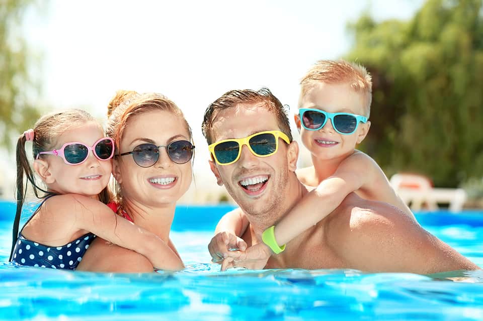 Family of four swimming in bright blue pool wearing colorful sunglasses