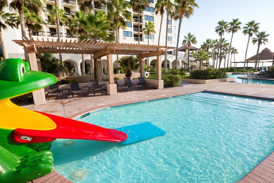 Resort pool with green water slide, pergola shade structure, palm trees, and high-rise building