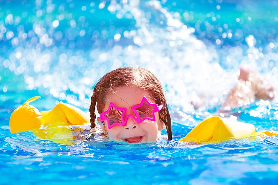 Young child swimming in bright blue pool wearing pink star-shaped sunglasses and yellow floaties