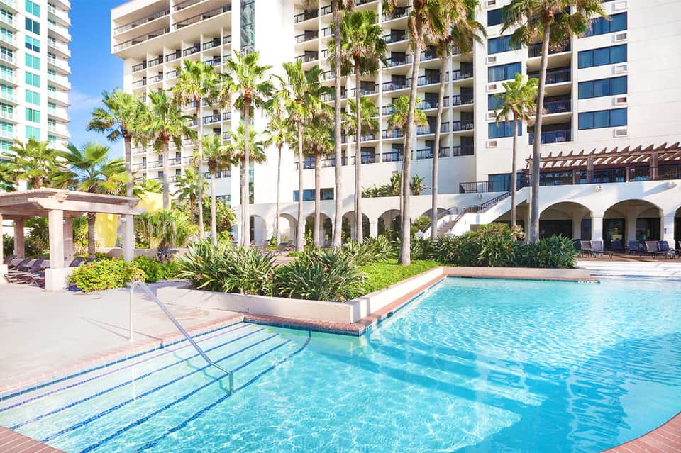 Resort pool surrounded by palm trees with multi-story white building and covered lounging areas