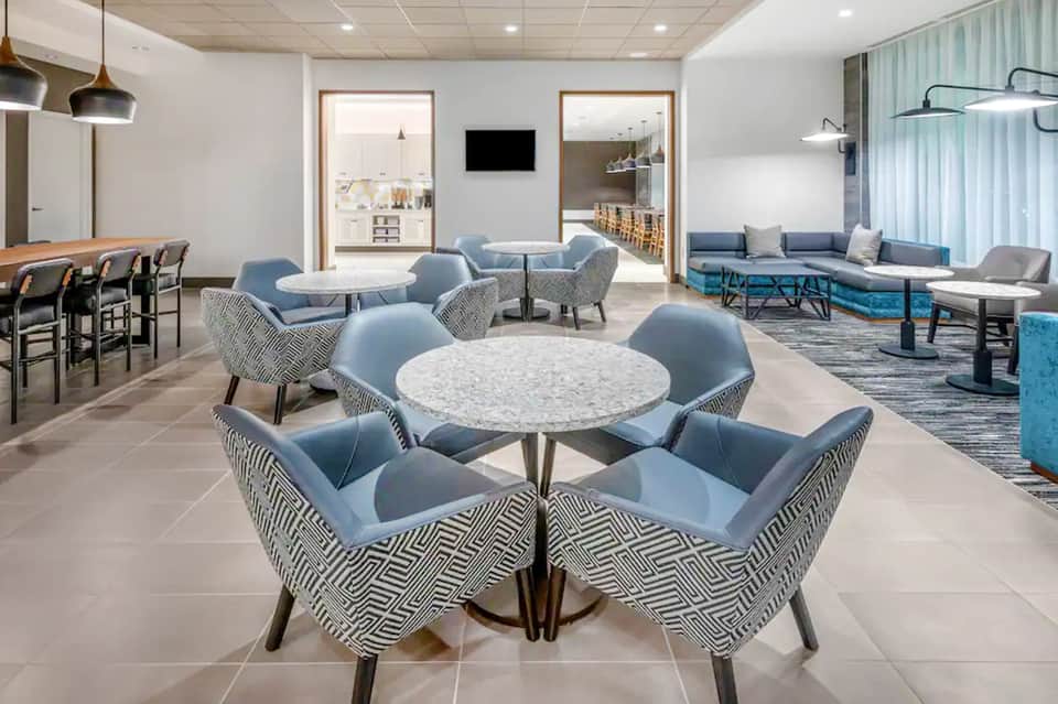 Modern hotel lobby with round tables, patterned chairs in blue, bar seating, and contemporary furnishings