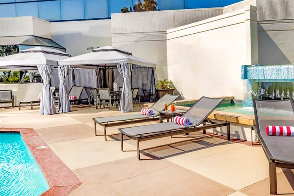 Luxury resort pool deck with lounge chairs, umbrellas, cabanas, and modern white building under clear blue sky