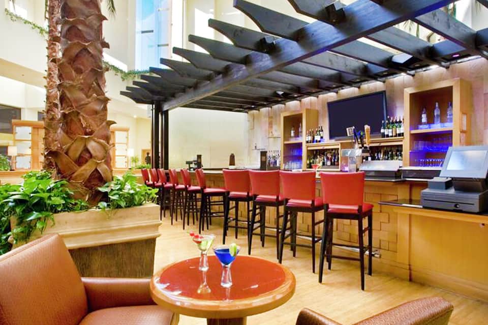Modern bar lounge with red accents, palm tree, open pergola, and illuminated beverage display