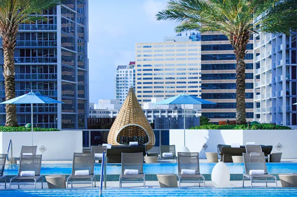 Luxury rooftop pool with lounge chairs, palm trees, and blue umbrellas overlooking city skyline
