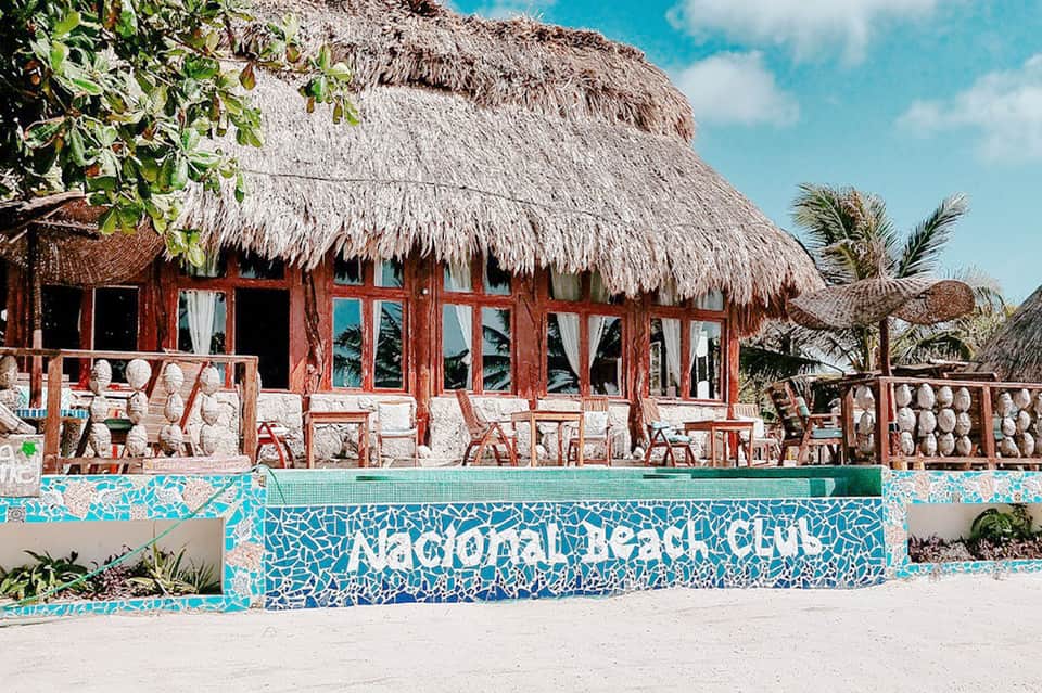 Colorful beachfront beach club with red wooden structure, thatched roof, and turquoise mosaic tile signage
