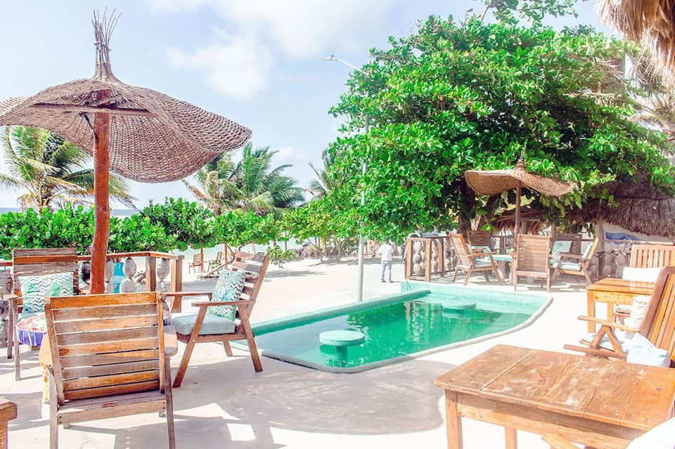 Tropical beach resort with small green pool, wooden loungers, and thatched umbrellas