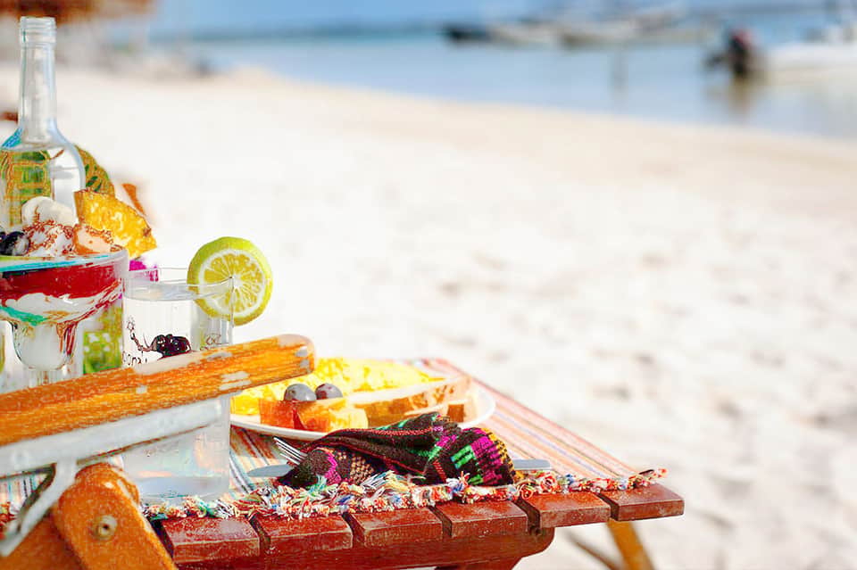 Beach picnic setup with wine bottle, cocktail with lime, fresh fruit and pastries on wooden table overlooking sandy beach