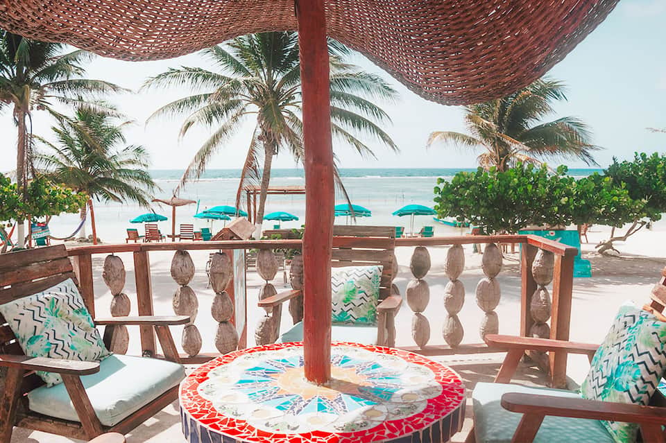 Beachfront lounge area with wooden railings, colorful round table, palm trees, and turquoise umbrellas overlooking the ocean