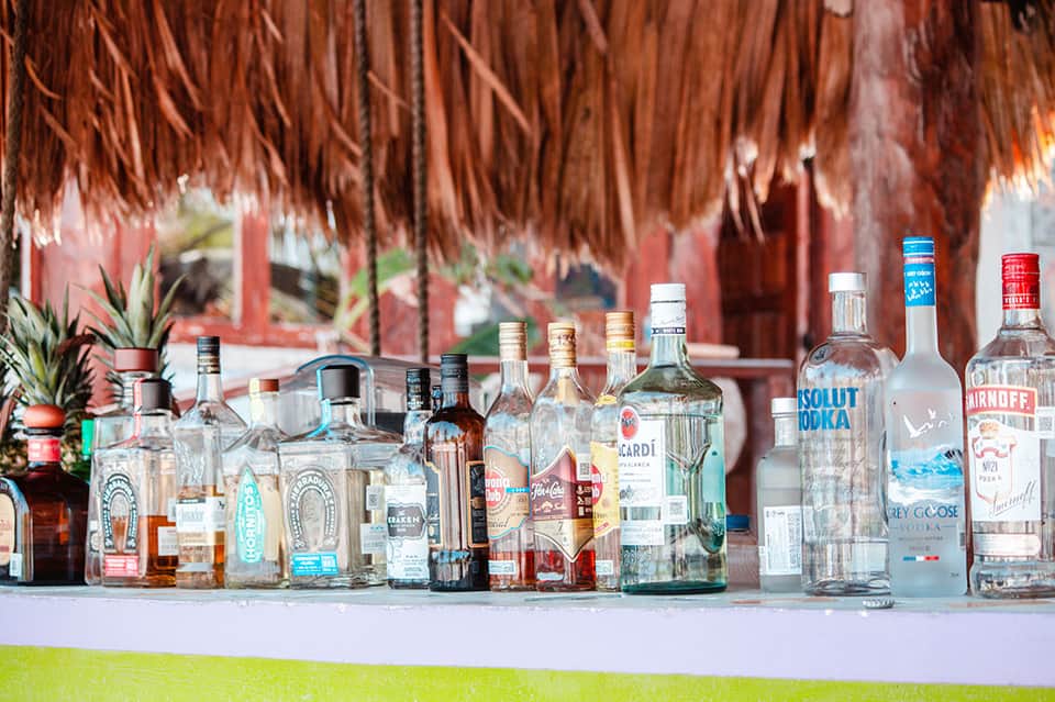 Array of liquor bottles displayed on a white table with tropical tiki bar in background