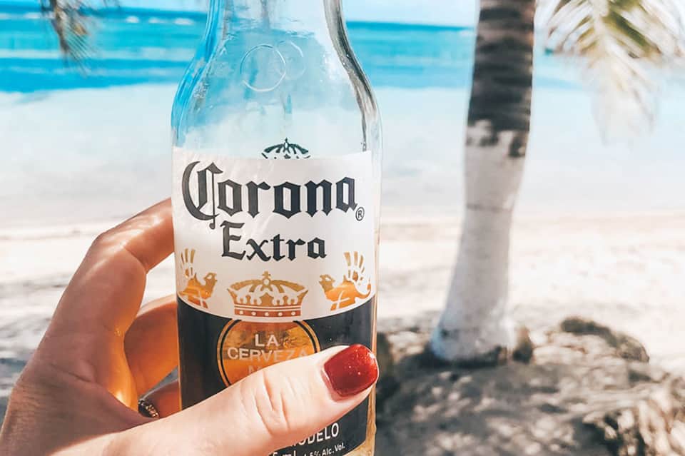 Corona Extra beer bottle held at beach with turquoise water and palm tree
