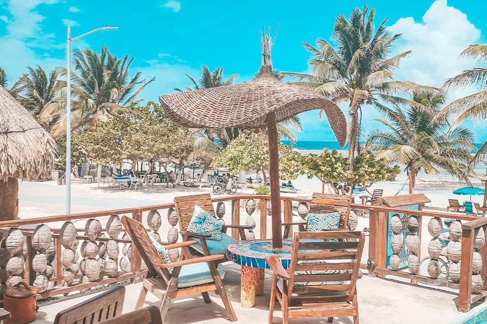 Tropical beach lounge with wooden chairs, thatched pergola, and palm trees