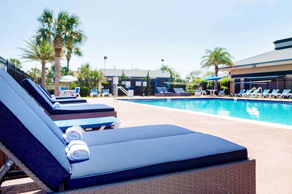 Resort pool deck with turquoise water, blue lounge chairs, palm trees, and covered cabanas in daytime