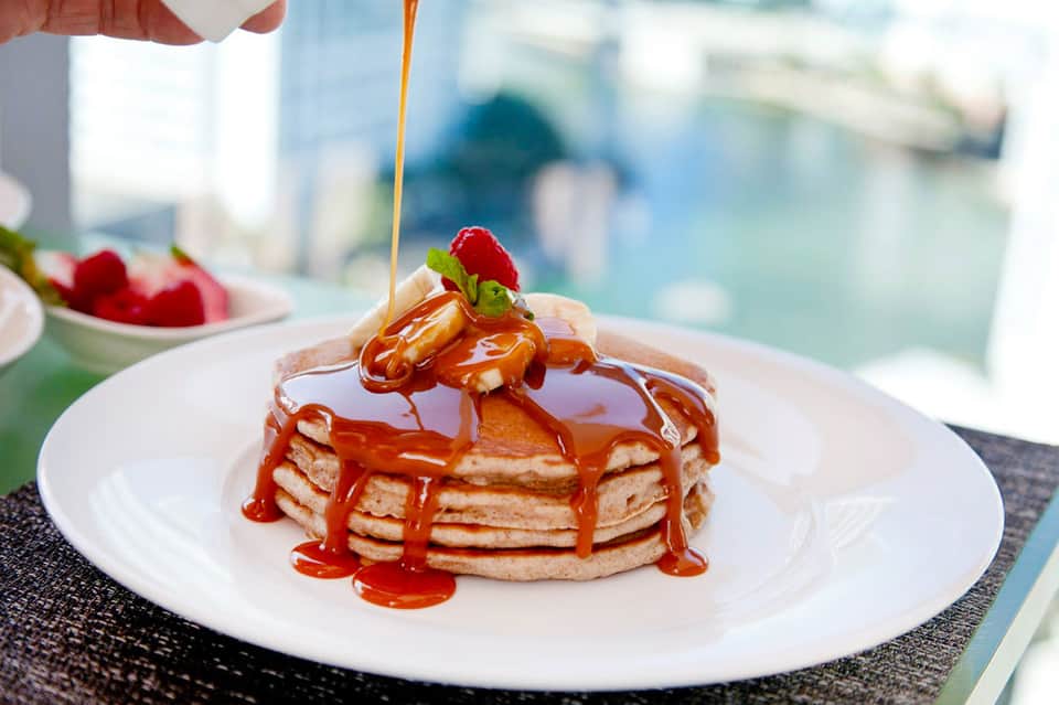 Stack of pancakes with maple syrup drizzle and fresh berries on white plate, waterfront view in background