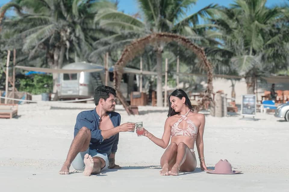 Couple toasting drinks on a sandy beach with palm trees and beach structures in the background