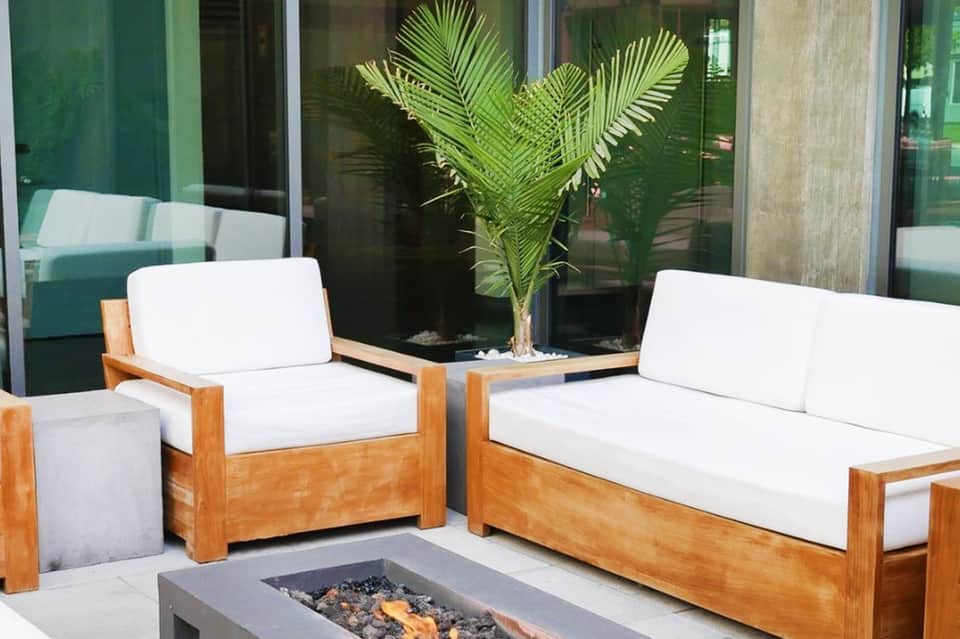 Modern outdoor lounge seating with white cushions, wooden frames, potted palm plant, and fire pit