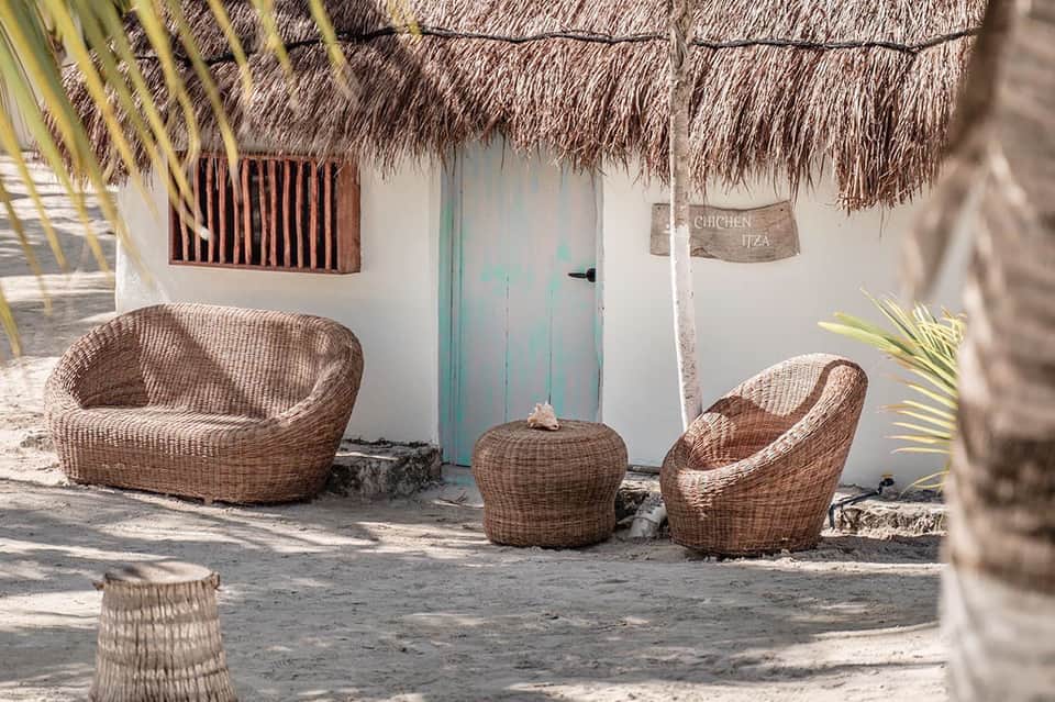 Woven rattan chairs on sand outside a white bungalow with thatched roof