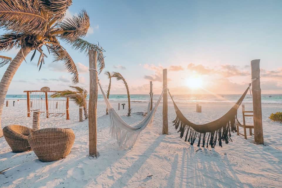 Beach sunset scene with hammocks, wooden swing, palm trees, and sandy shore at golden hour