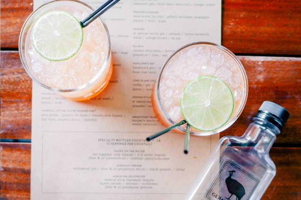 Two tropical cocktails with lime slices and black straws on wooden table next to menu and bottle