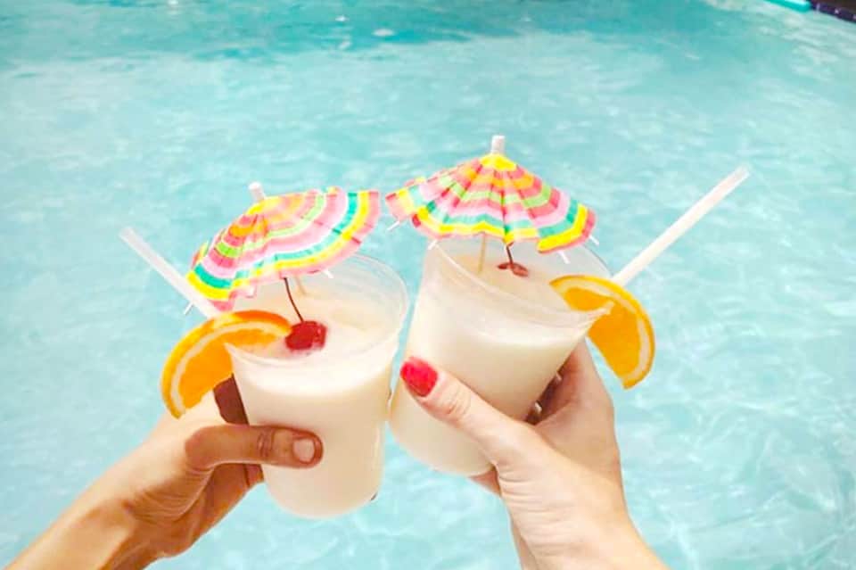 Two tropical cocktails with colorful umbrellas held above a swimming pool