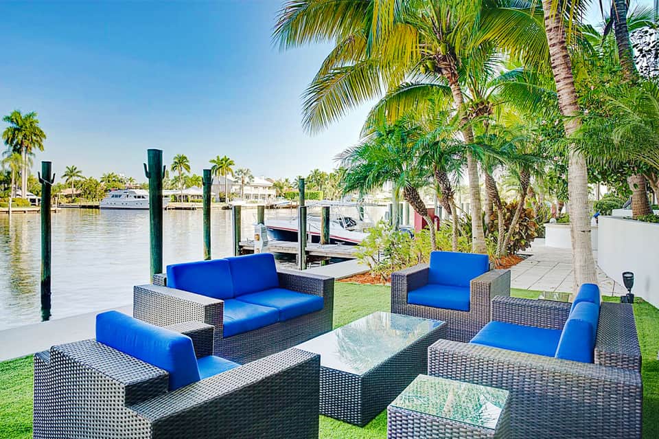 Waterfront lounge area with blue cushioned furniture, palm trees, docked boats, and manicured lawn