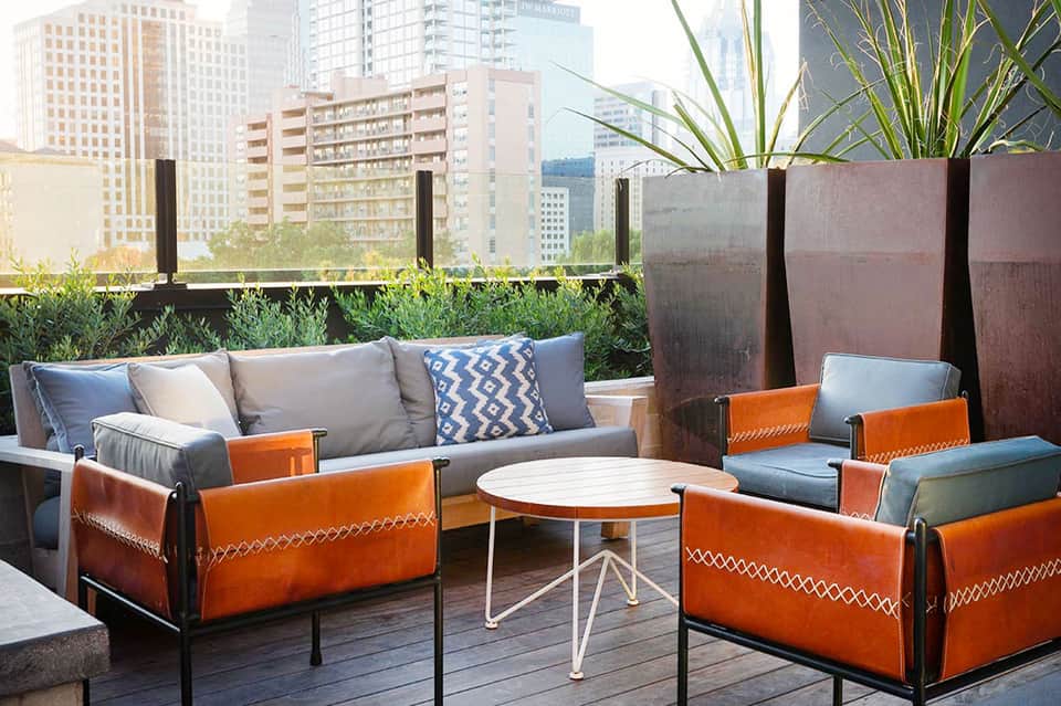 Rooftop lounge with orange leather chairs, gray sofa, white table, and city skyline view with potted plants