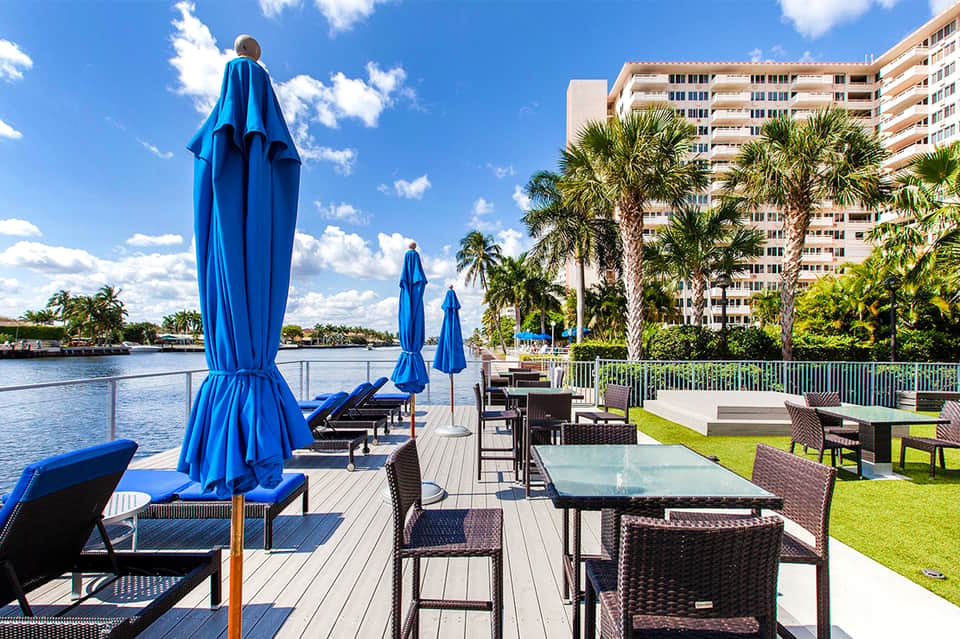 Waterfront resort deck with blue umbrellas, lounge chairs, and palm trees overlooking the water