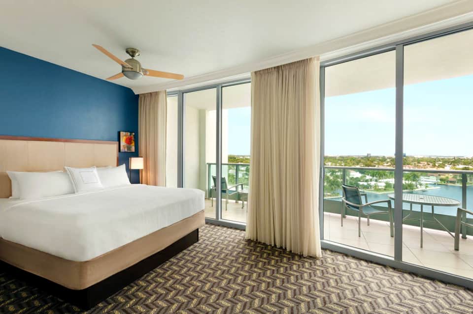 Modern hotel bedroom with blue accent wall, balcony overlooking water and trees