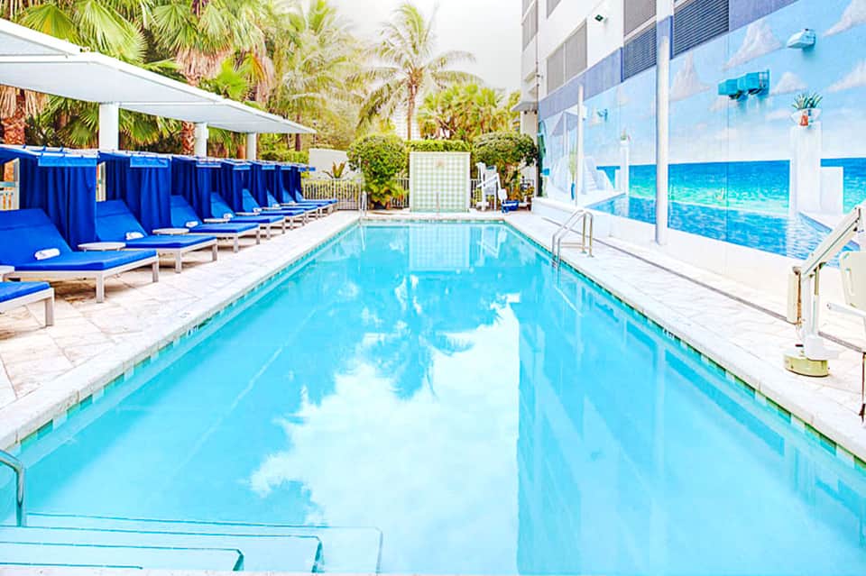 Indoor lap pool with blue water, palm trees, blue cabanas, and large windows overlooking waterfront