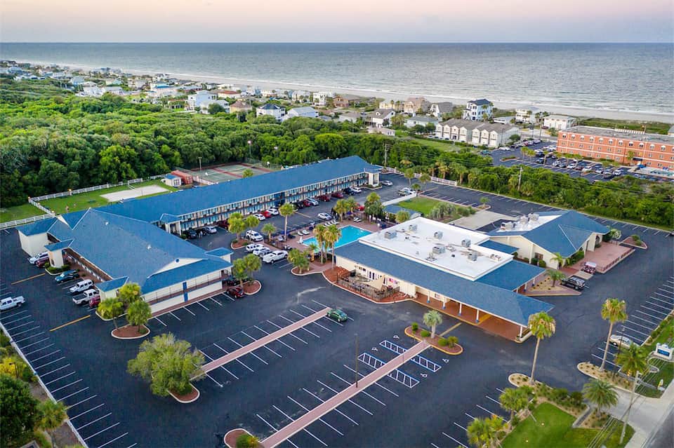 Aerial view of beachfront resort with blue roof buildings, pool, parking, and ocean backdrop