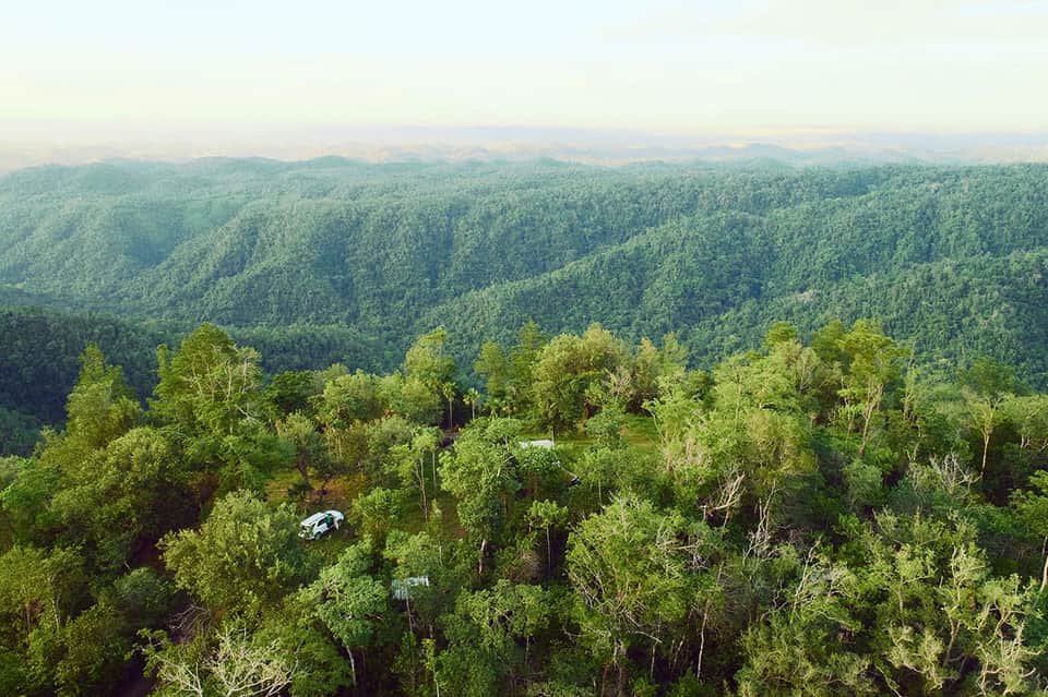 Aerial view of lush green forested hills and valleys with white vehicle visible on a road