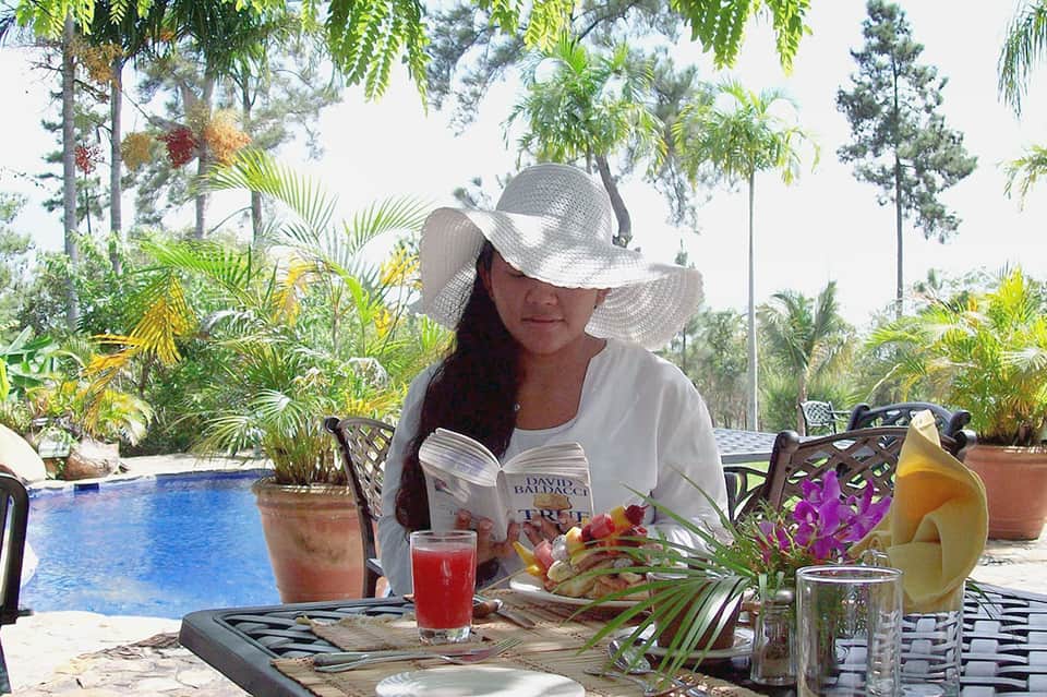 Woman reading poolside in tropical setting with fresh beverages, flowers, and lush garden landscape