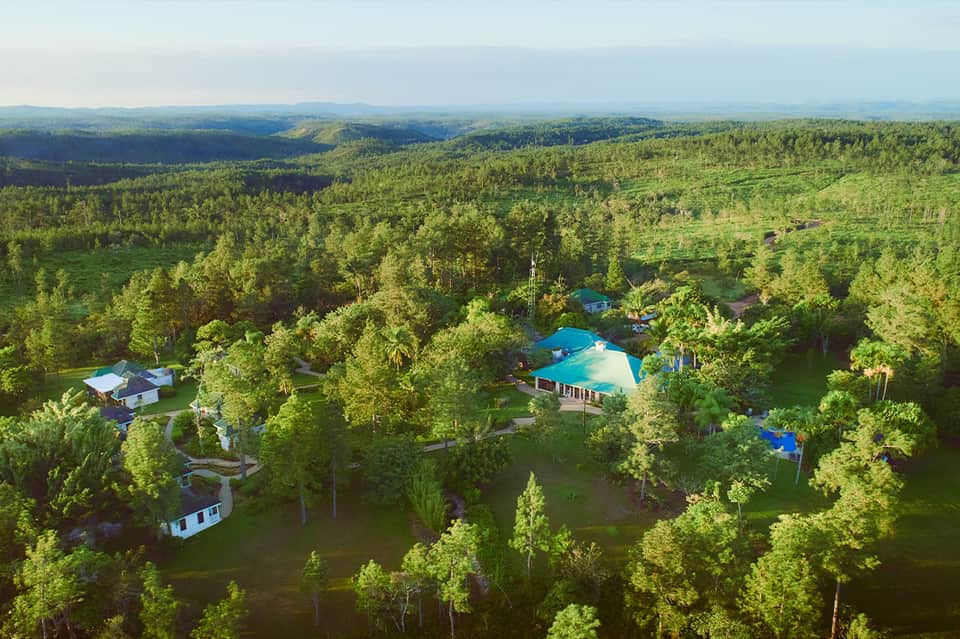Aerial view of resort pools and buildings nestled in dense green forest with mountains in distant background