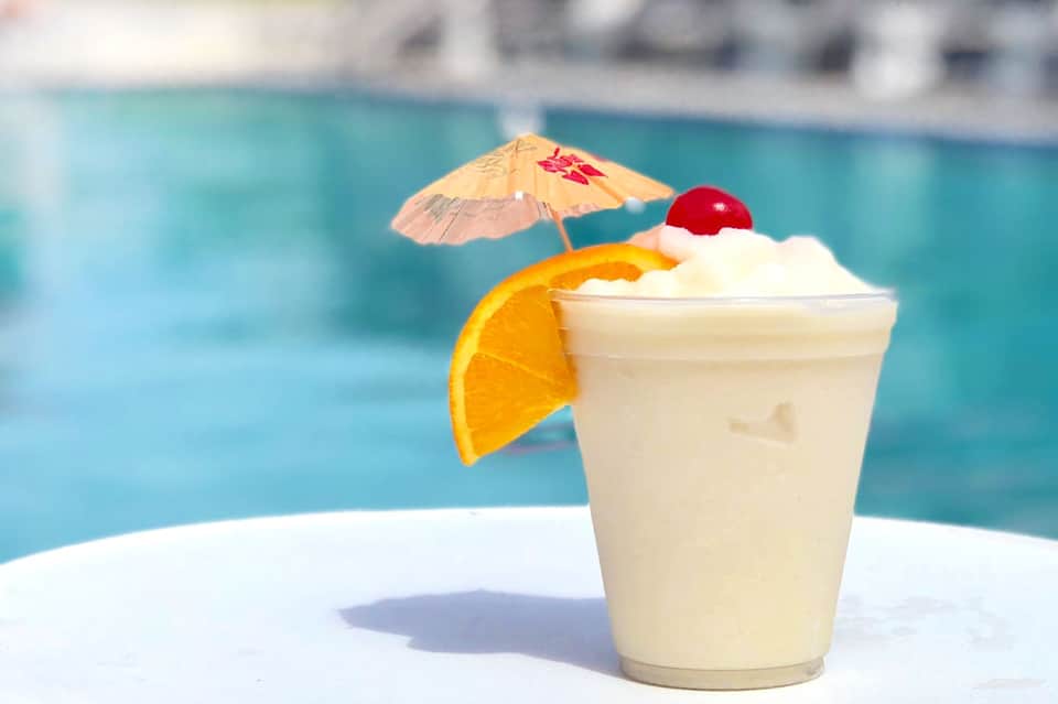 Tropical cocktail with orange slice and umbrella garnish sitting poolside with turquoise water in background