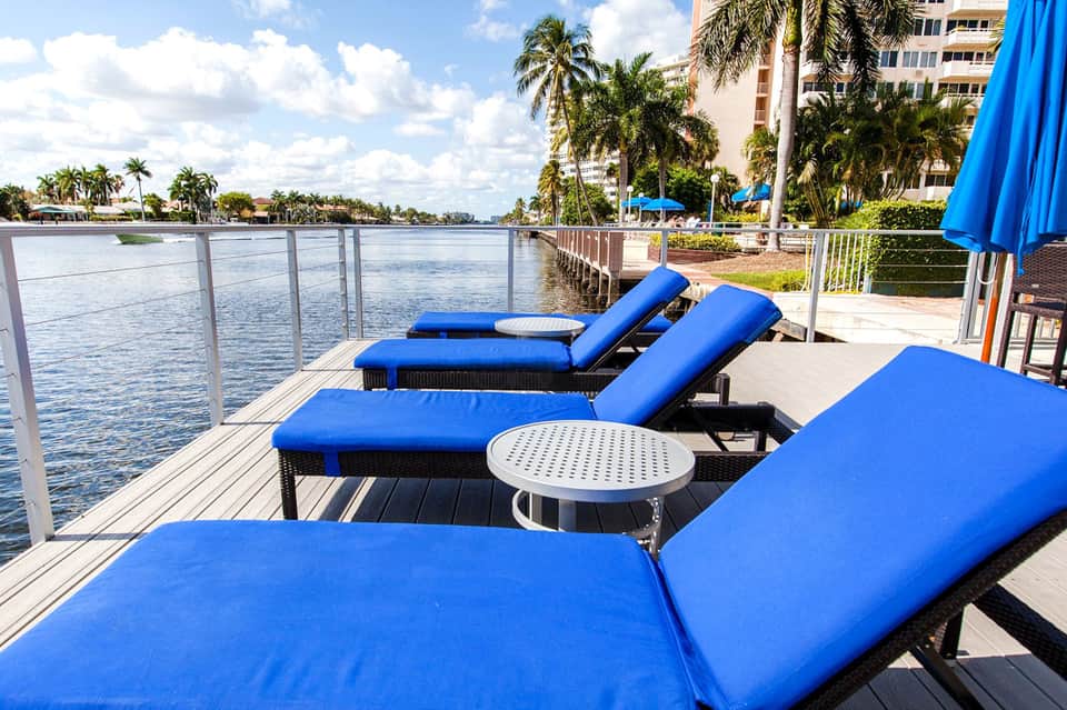 Waterfront lounge area with blue chaise lounges overlooking canal and palm trees