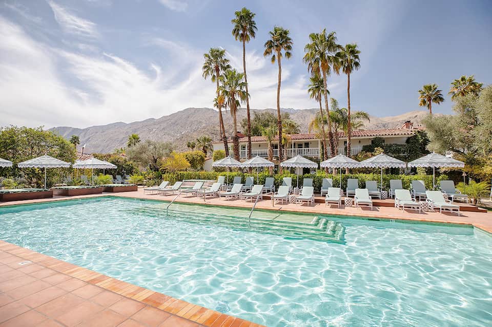 Resort pool with palm trees, white umbrellas, lounge chairs, and Spanish colonial-style building with mountains