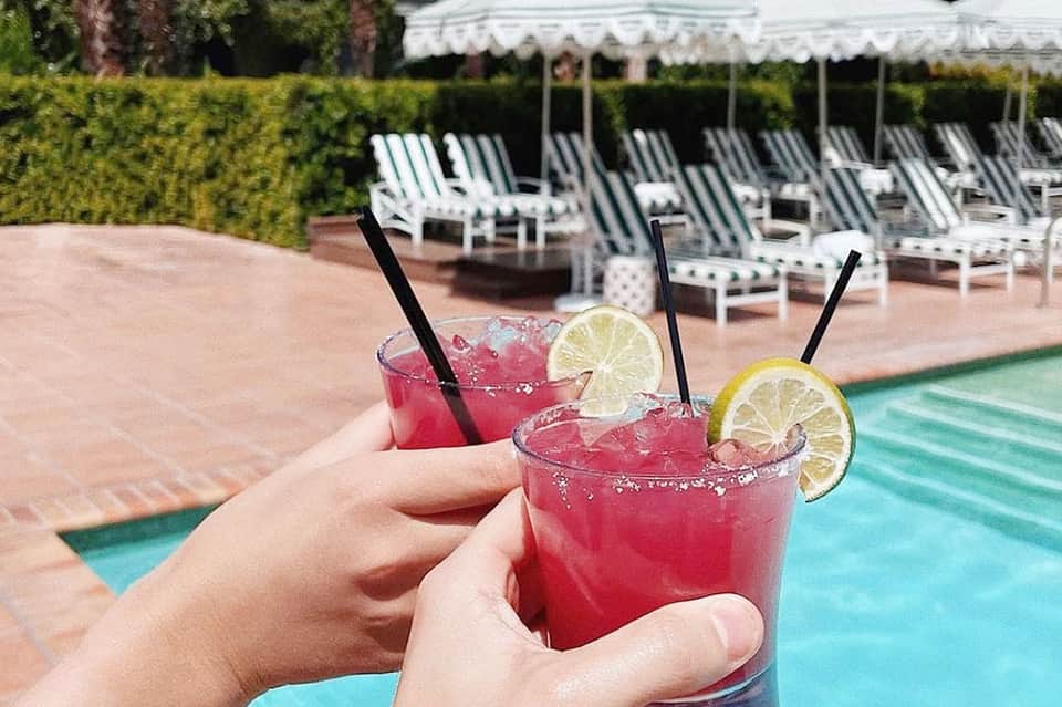 Hands holding pink cocktails with lime garnish poolside with striped beach chairs and umbrellas in background