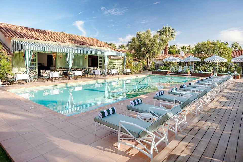 Resort pool with white lounge chairs, striped cabana, tropical landscaping, and palm trees under blue sky