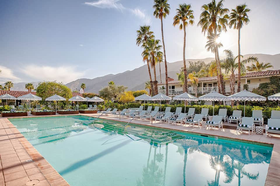 Resort pool with white umbrellas, palm trees, and mountain views at sunset