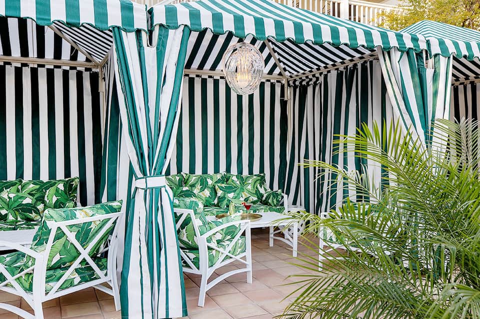 Striped cabana with green and white awning, white lounge chairs, and tropical plants on patio