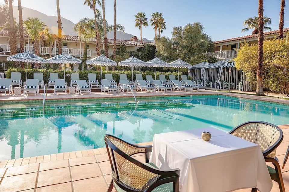 Resort pool with lounge chairs under umbrellas, palm trees, and mountain views