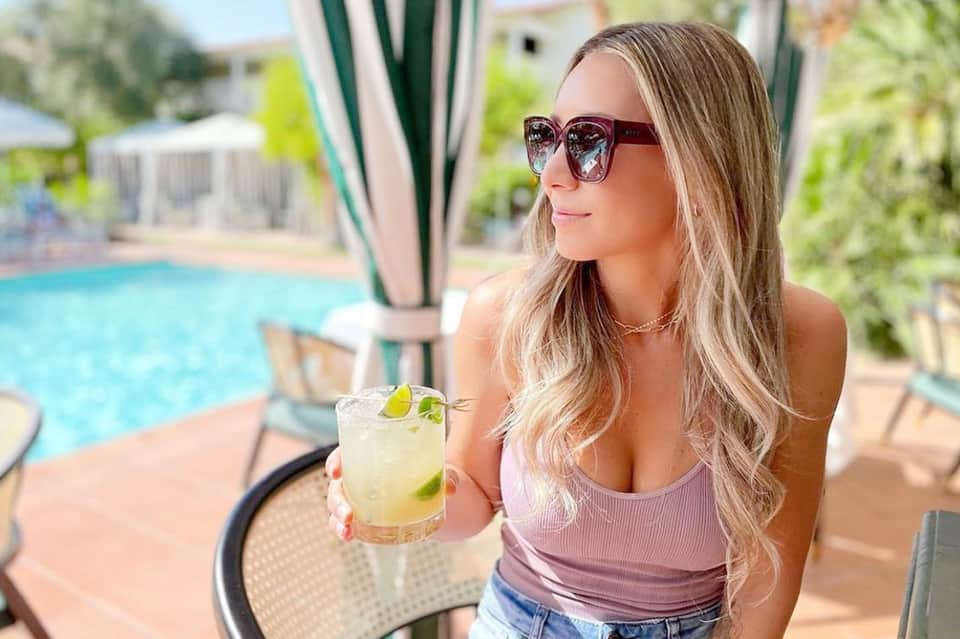 Woman enjoying cocktail by resort pool with lounge chairs and tropical landscaping visible