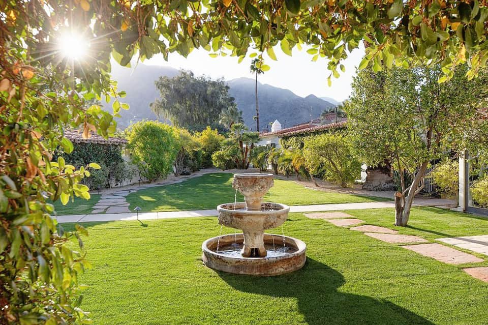 Lush resort garden courtyard with fountain, manicured lawn, mature trees, and mountain backdrop